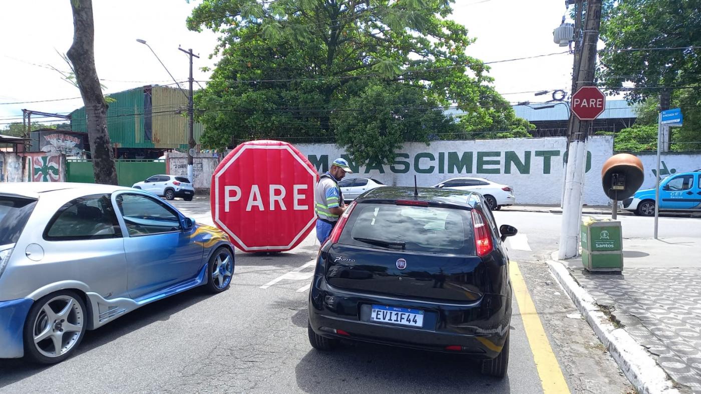 agente da cet está orientando motorista dentro de carro. Na via, à frente, há um grande inflável onde se lê Pare. #paratodosverem