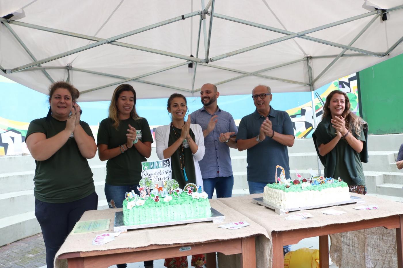 Quatro mulheres e dois homens batem palmas atrás de mesa com bolo de aniversário na cor verde