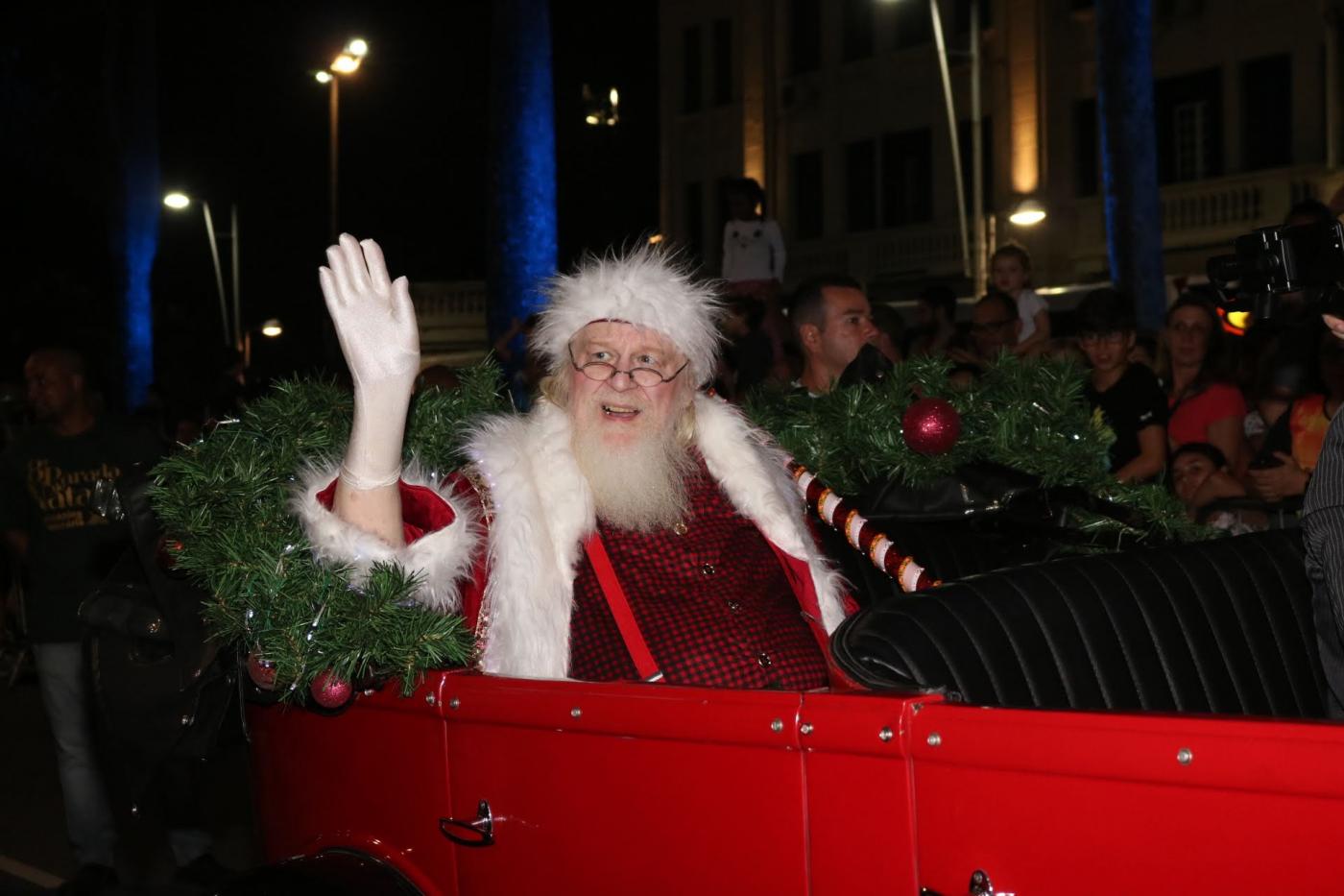 Idoso vestido de papai noel acena ao público em banco traseiro de carro