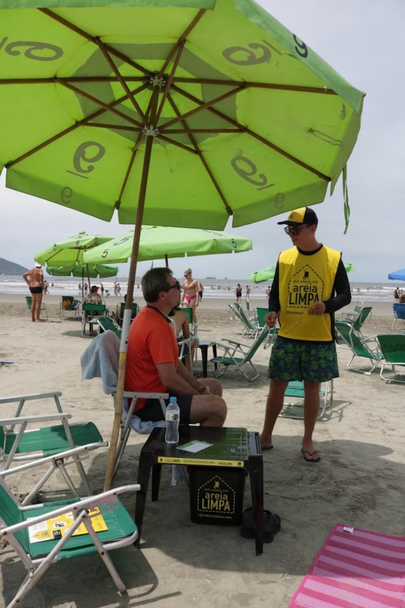Agente uniformizado conversa com banhista que está sentado em cadeira na praia. Ao lado há apetrechos para o descarte correto de lixo. #Pracegover