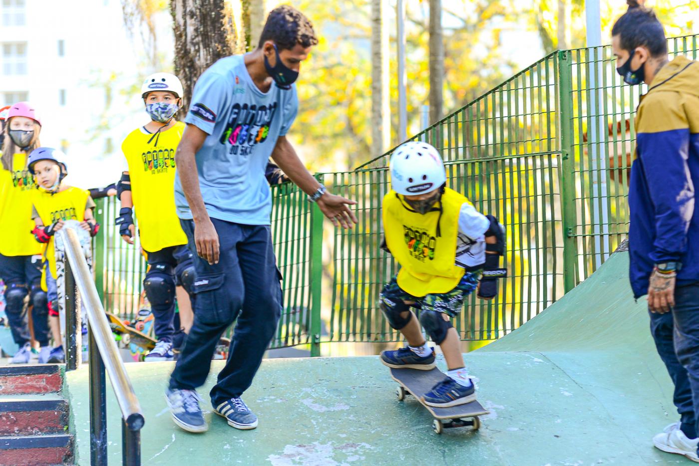 Criança andando de skate desce rampa sendo orientada por professor. #paratodosverem