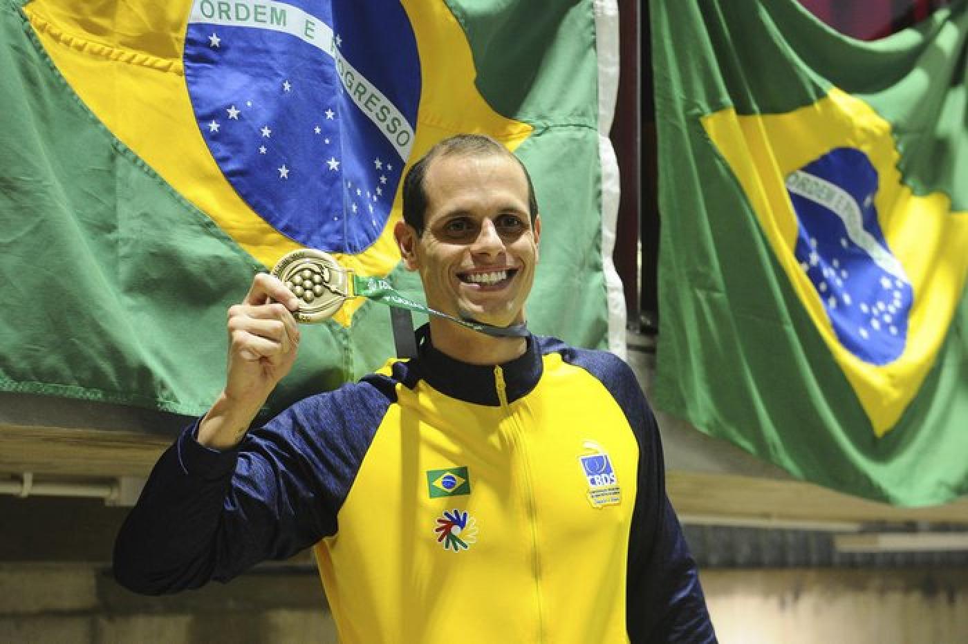 atleta exibe medalha com bandeira do brasil ao fundo #paratodosverem