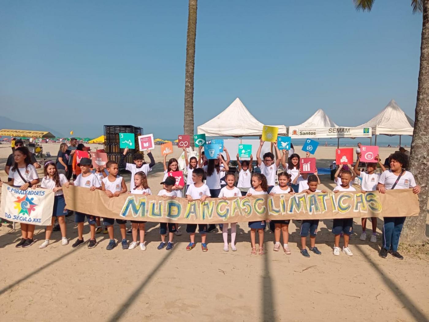 alunos posam para foto na faixa de areia da praia segurando uma faixa onde se mudanças climáticas. #paratodosverem 