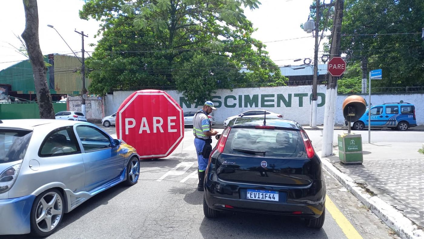 agente da CET na rua está abordando motorista que está dentro de carro. Ao fundo, um grande inflável onde se lê Pare. #paratodosverem