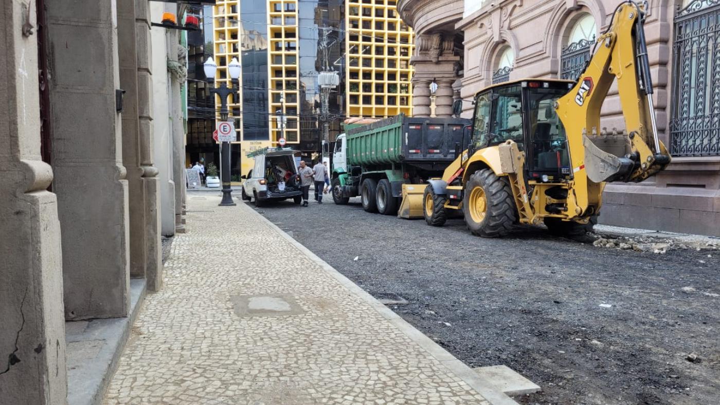 rua em obras com calçada à esquerda já coberta por pedras portuguesas. Há um trator na via. #paratodosverem 