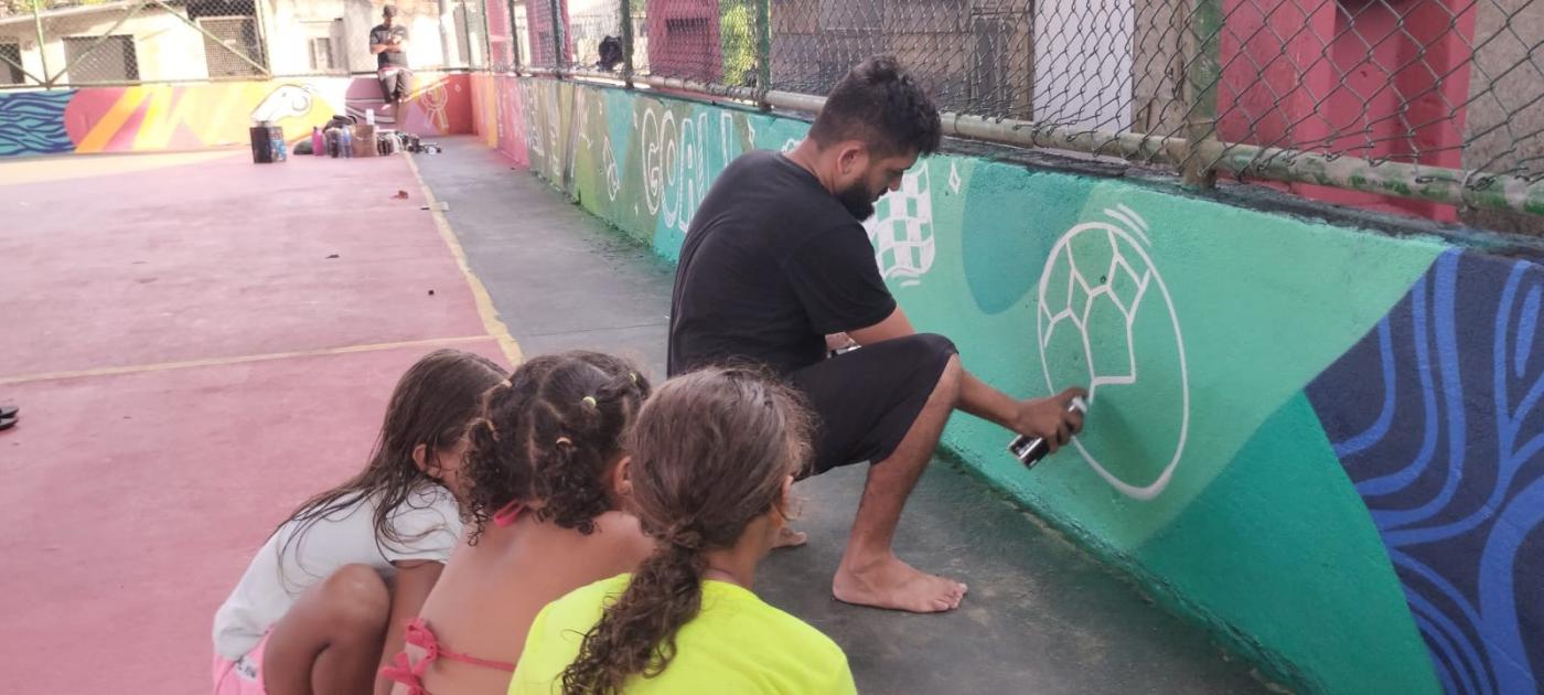 artista desenha e pinta bola no muro de quadra. Crianças, de costas para a foto, observam os movimentos do artista. #paratodosverem