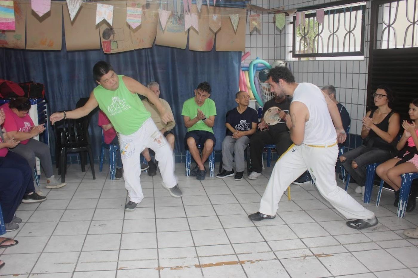 jovens fazem  movimentos de capoeira diante de roda de pessoas sentadas. #paratodosverem 