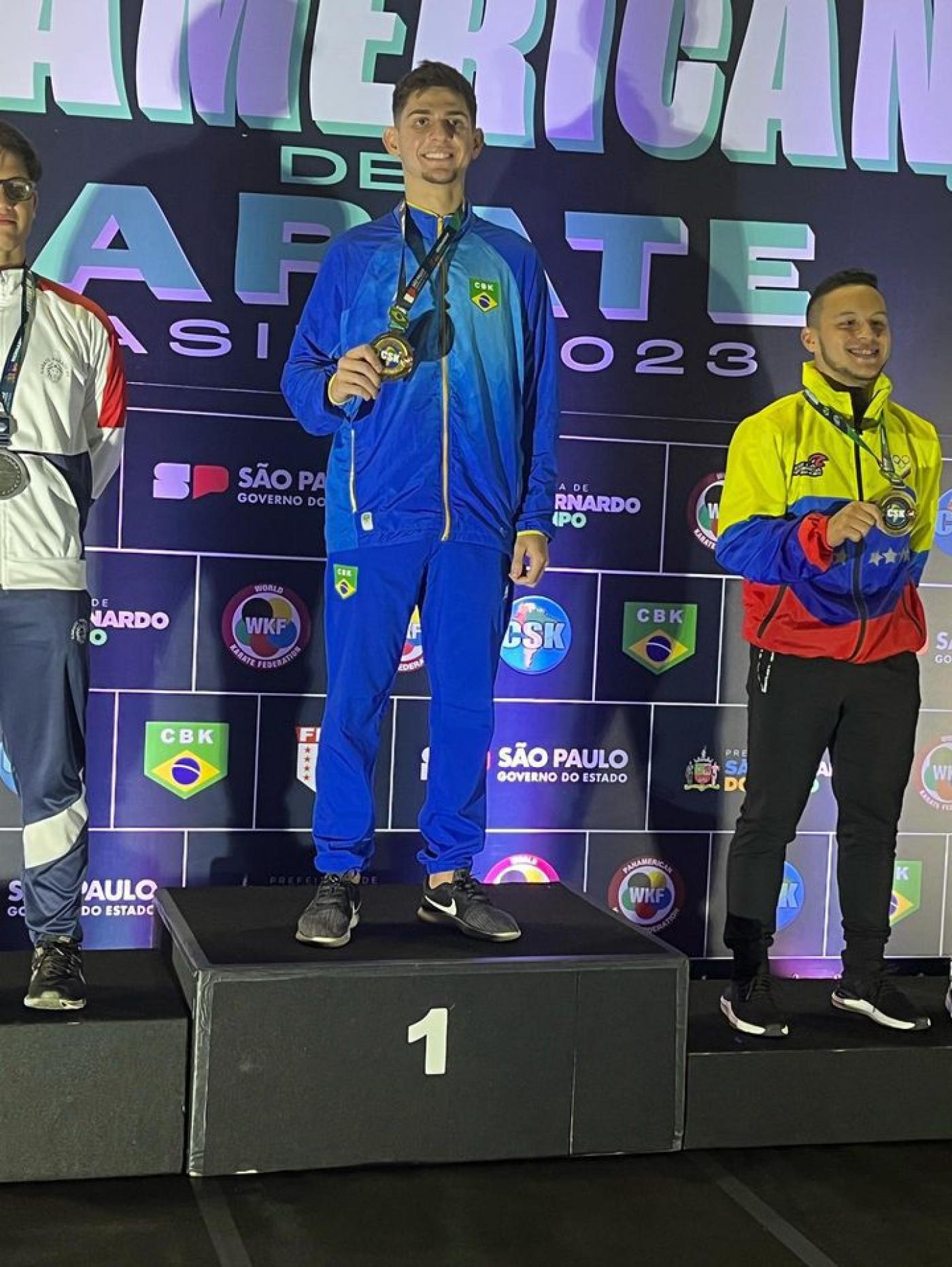 matheus está no primeiro lugar do pódio com a medalha no peito e a mão direita na medalha. #paratodosverem