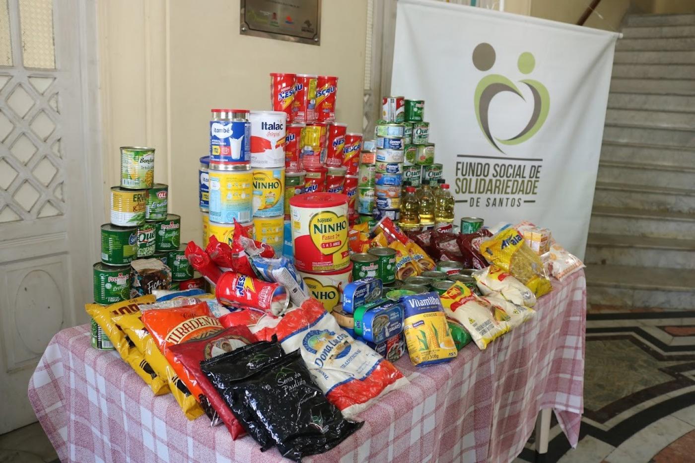 mesa com alimentos não perecíveis empilhados.Ao fundo, um banner do fundo social de solidariedade. #paratodosverem