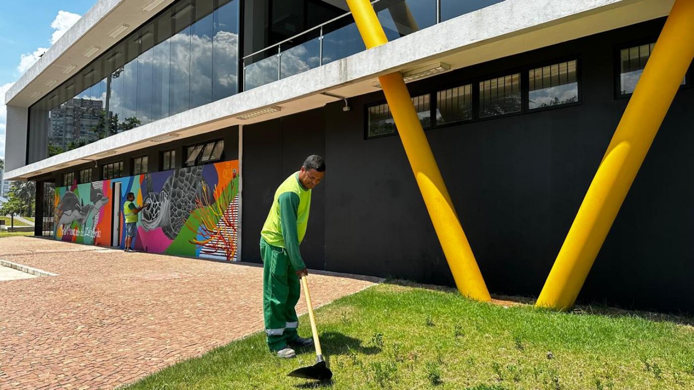 homem usa pá em gramado diante de fachada de prédio. #paratodosverem 