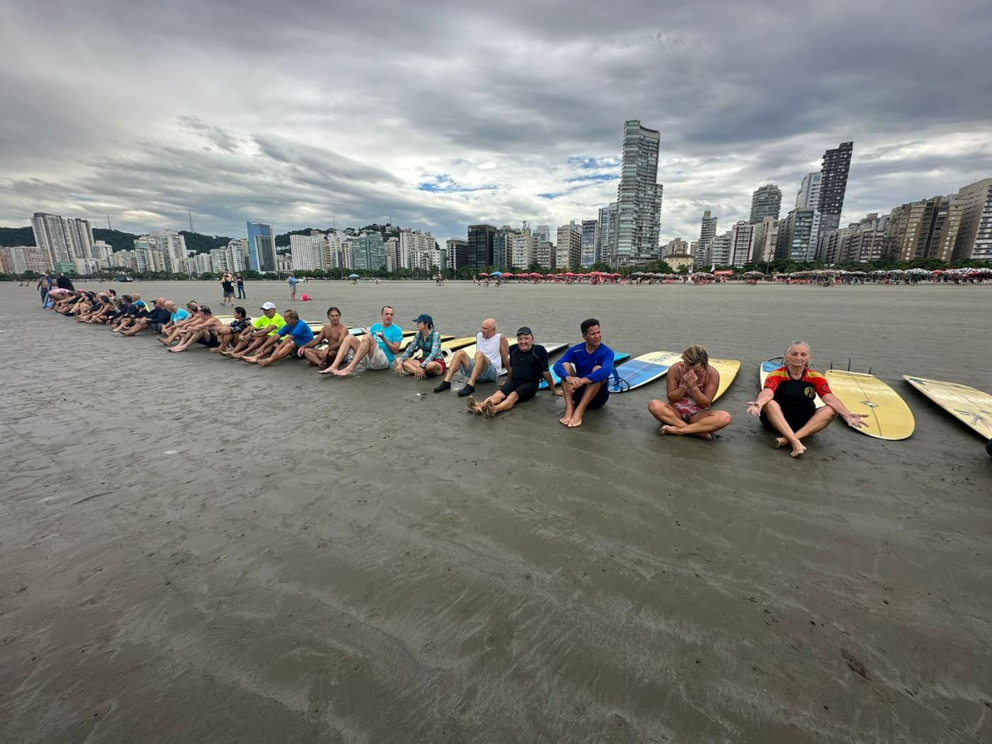 alunos estão sentados na areia em aquecimento para a aula. todos estão com pranchas. #paratodosverem