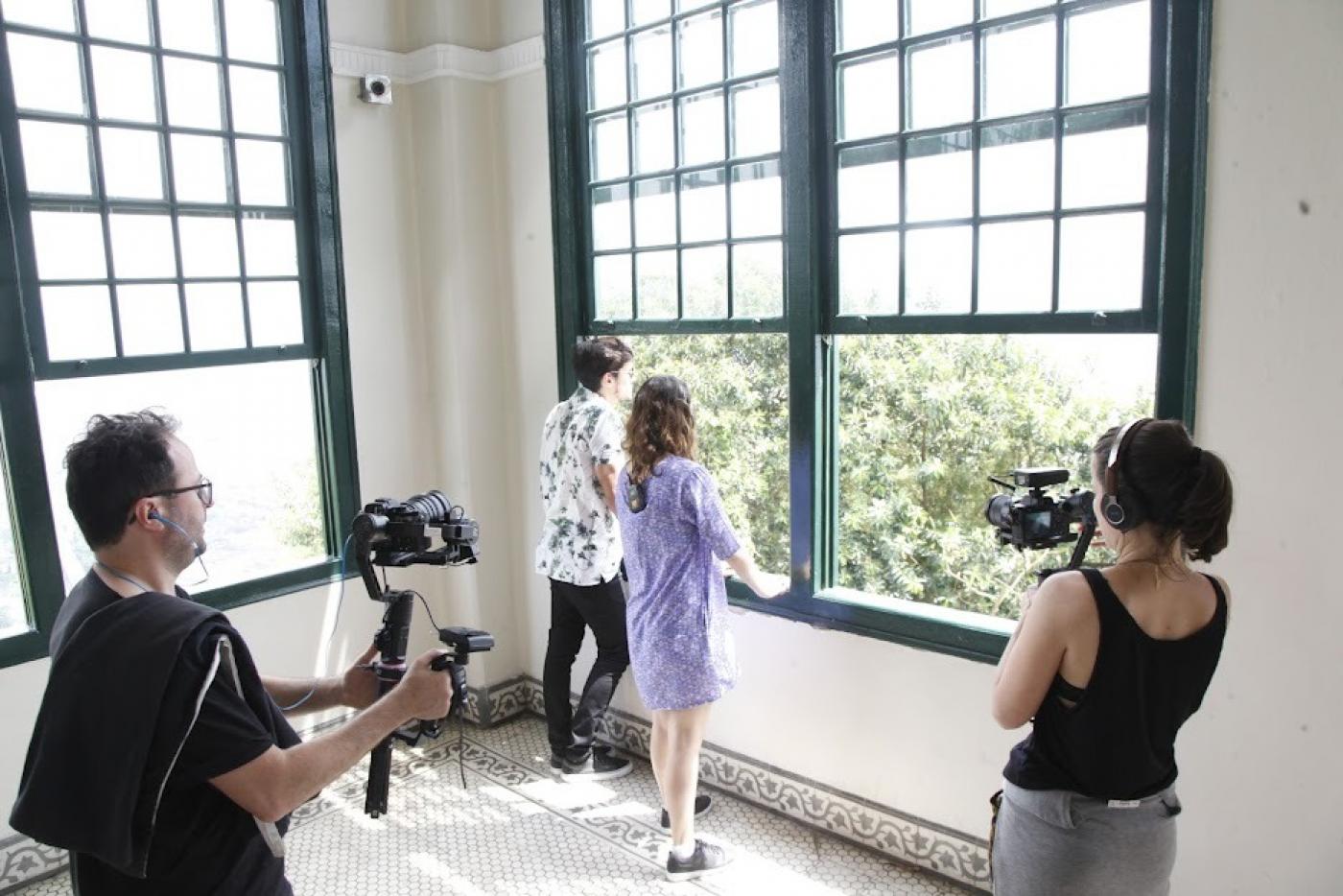 Duas pessoas filmam um casal encostado na janela olhando para fora #paratodosverem