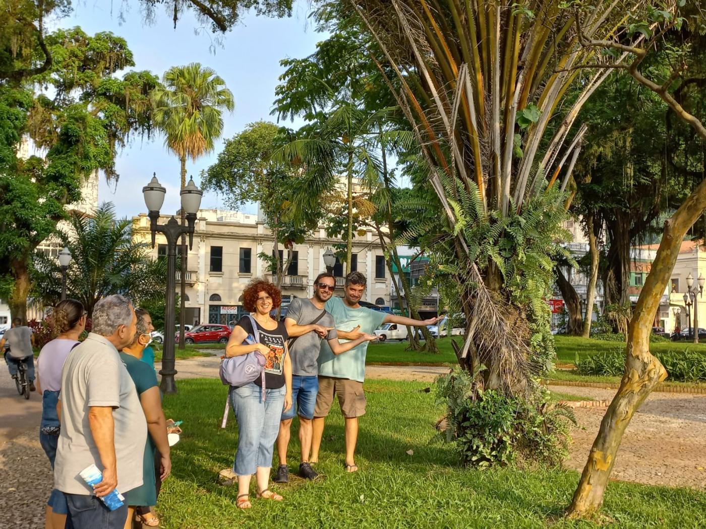 grupo de pessoas está em praça sobre a grama e com árvores em volta. #paratodosverem