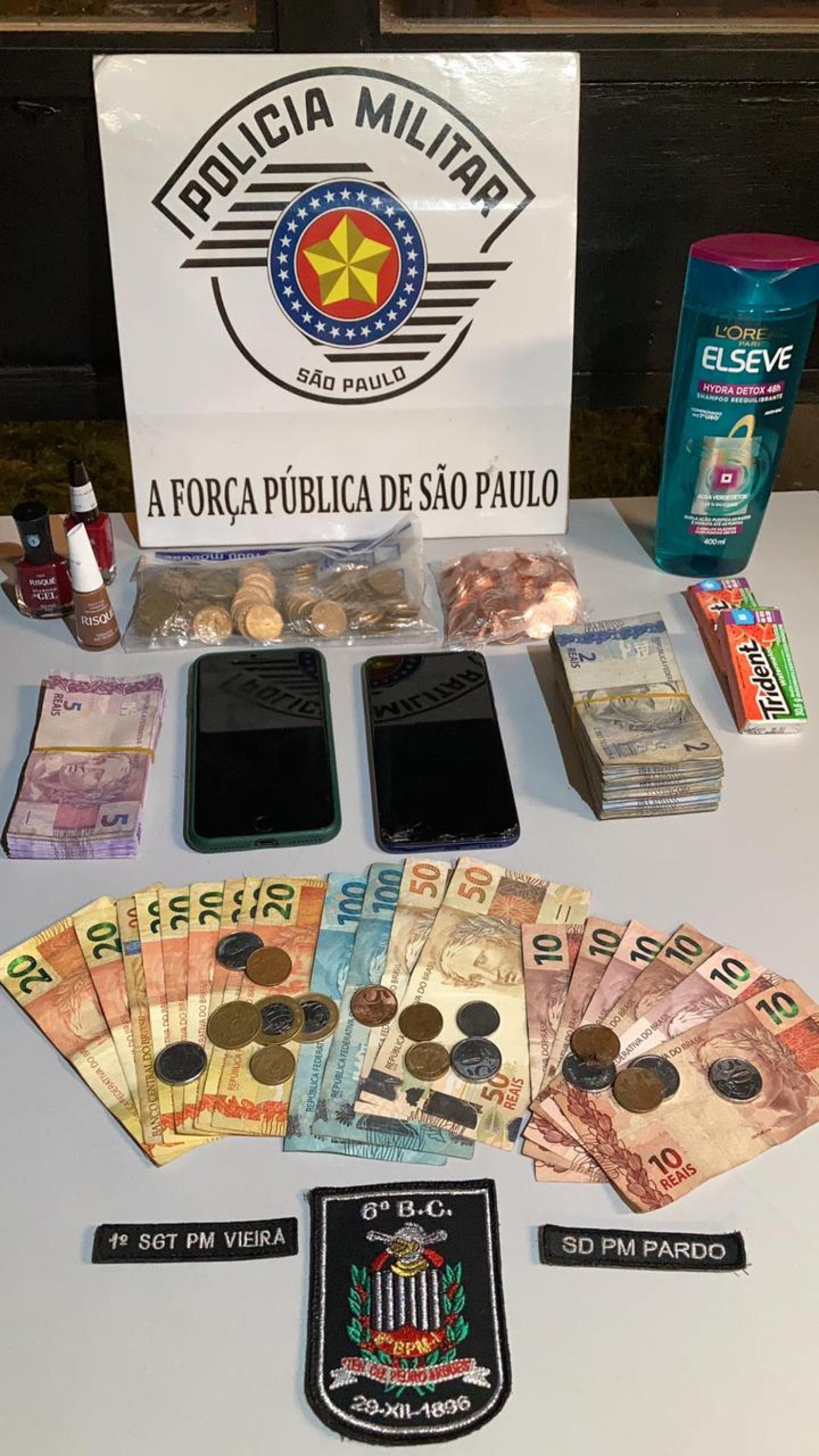 Mesa com cédulas de dinheiro, moedas, celulares e um shampoo. #paratodosverem