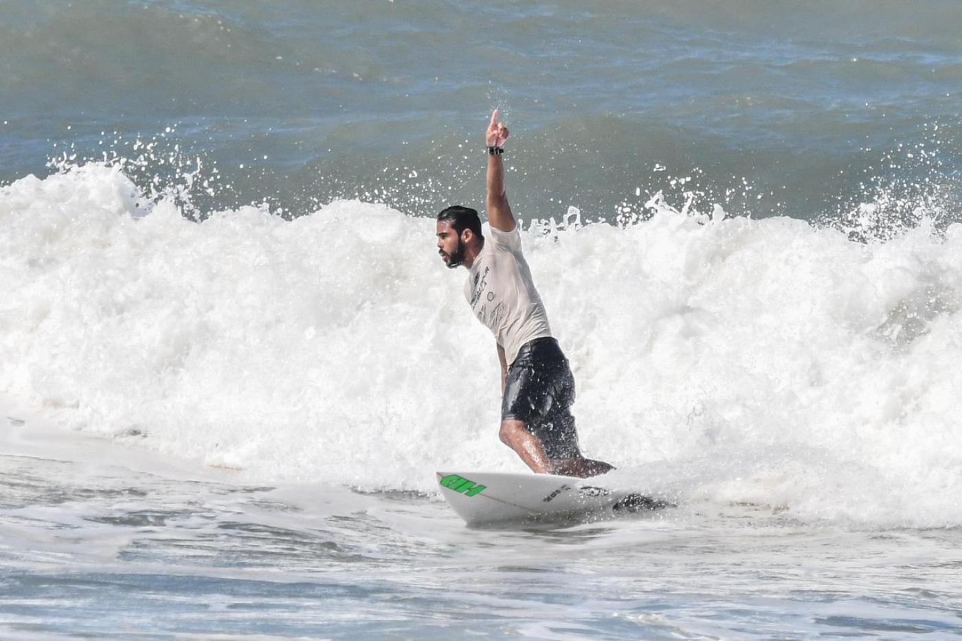 surfista está sobre a prancha no mar. Seu braço esquerdo está para o alto. #paratodosverem 