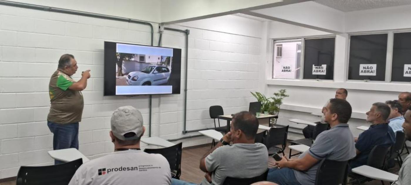 homem está em pé ao lado de um telão dando palestra para pessoas sentadas num pequeno auditório. #paratodosverem