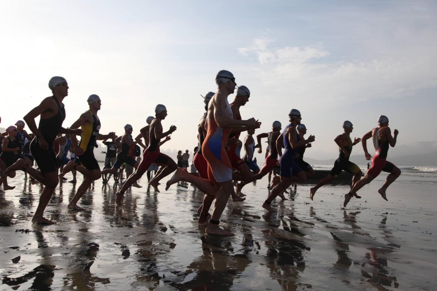 Atletas correm em direção a água na praia. #paratodosverem