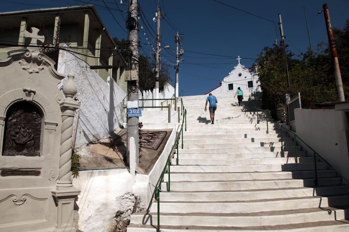 vista de nicho da Via Crucis à esquerda e trecho de escadaria com duas pessoas subindo. No alto, a capela. #paratodosverem