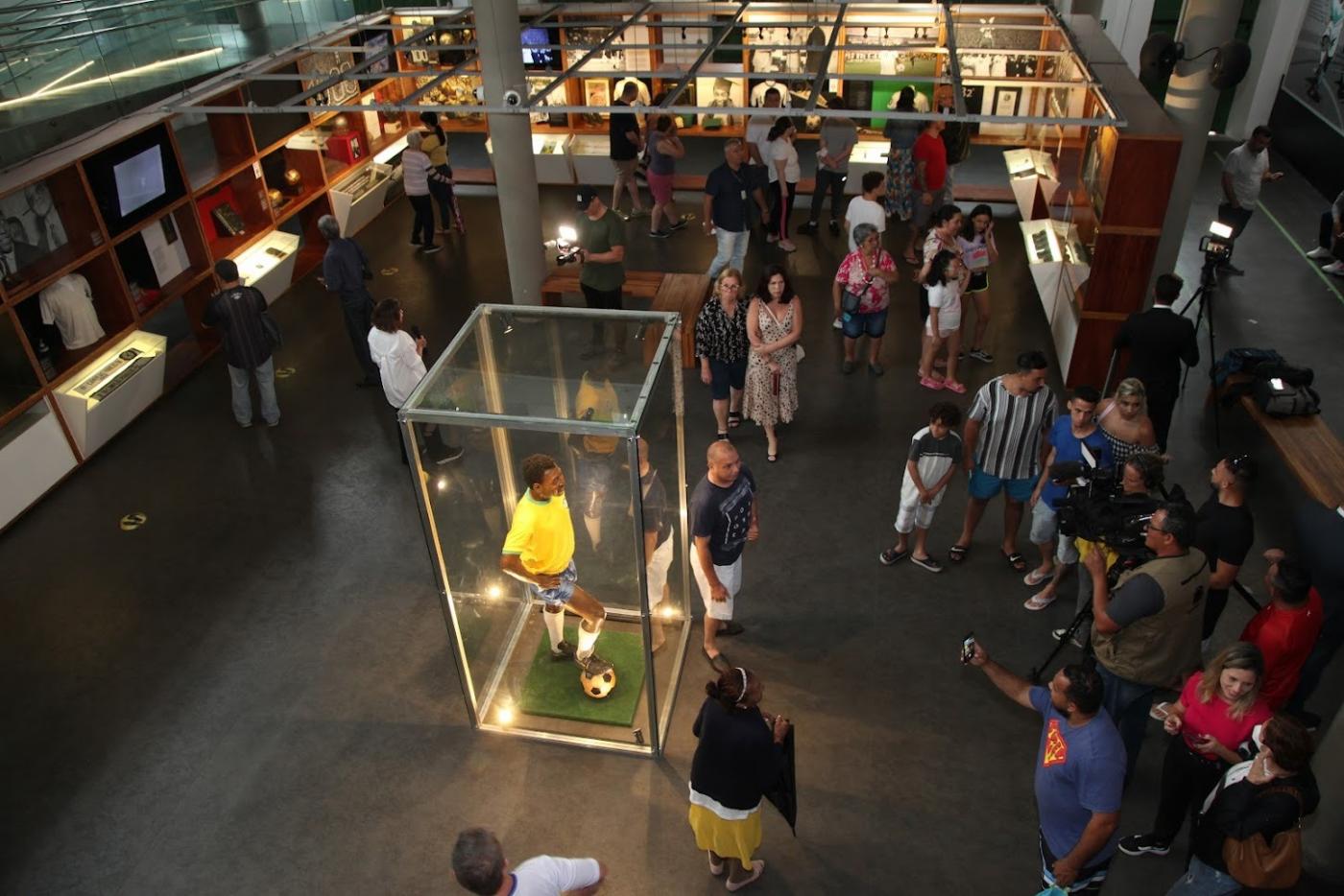 escultura de pelé dentro de redoma de vidro e vários visitantes circulando dentro de museu. #paratodosverem