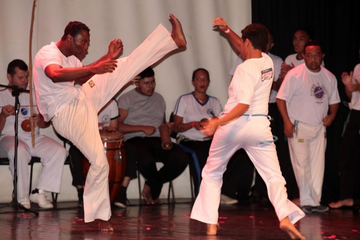 dois homens jogam capoeira. Músicos estão ao fundo. #paratodosverem