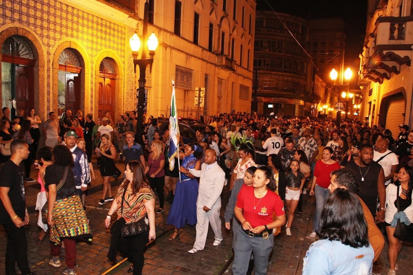 grande volume de pessoas caminha por rua do centro histórico à noite. Os postes de modelo republicano estão acesos. #paratodosverem 