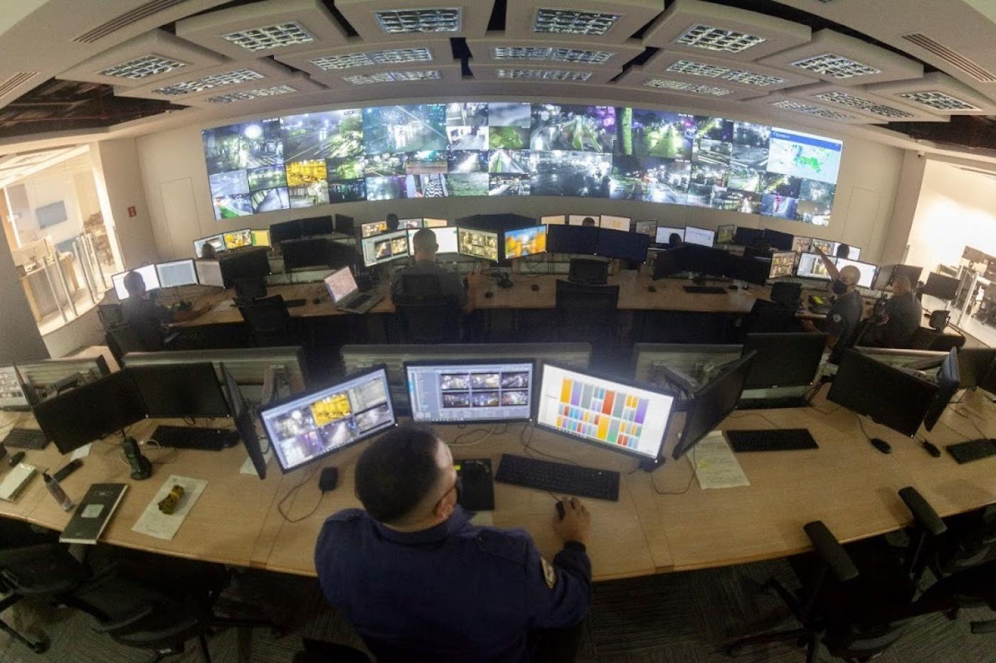 vista geral pelo alto do Centro de controle operacional, com um homem operando três computadores em primeiro plano e várias telas ao fundo. #paratodosverem