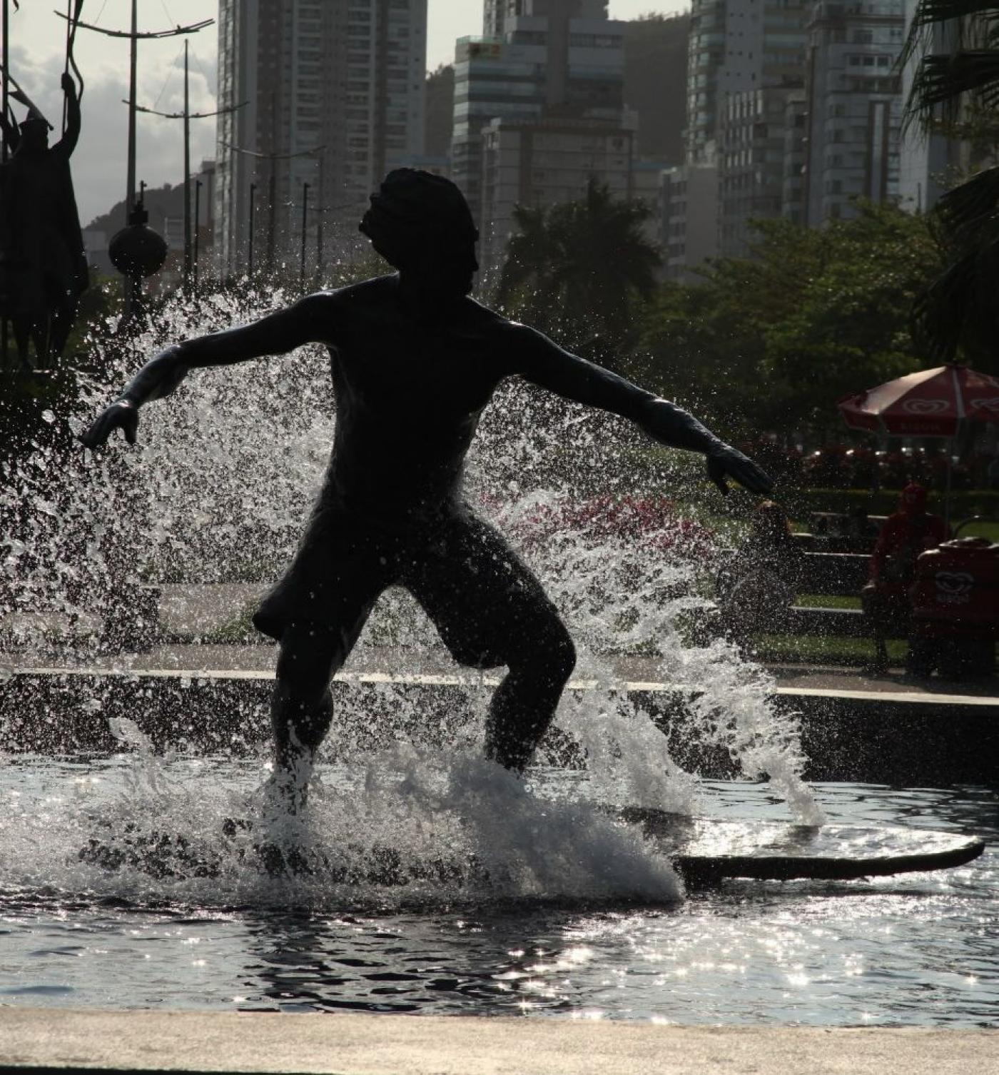 estátua do surfista contra a luz, parecendo uma sombra, com água jorrando sobre o atleta. #paratodosverem 