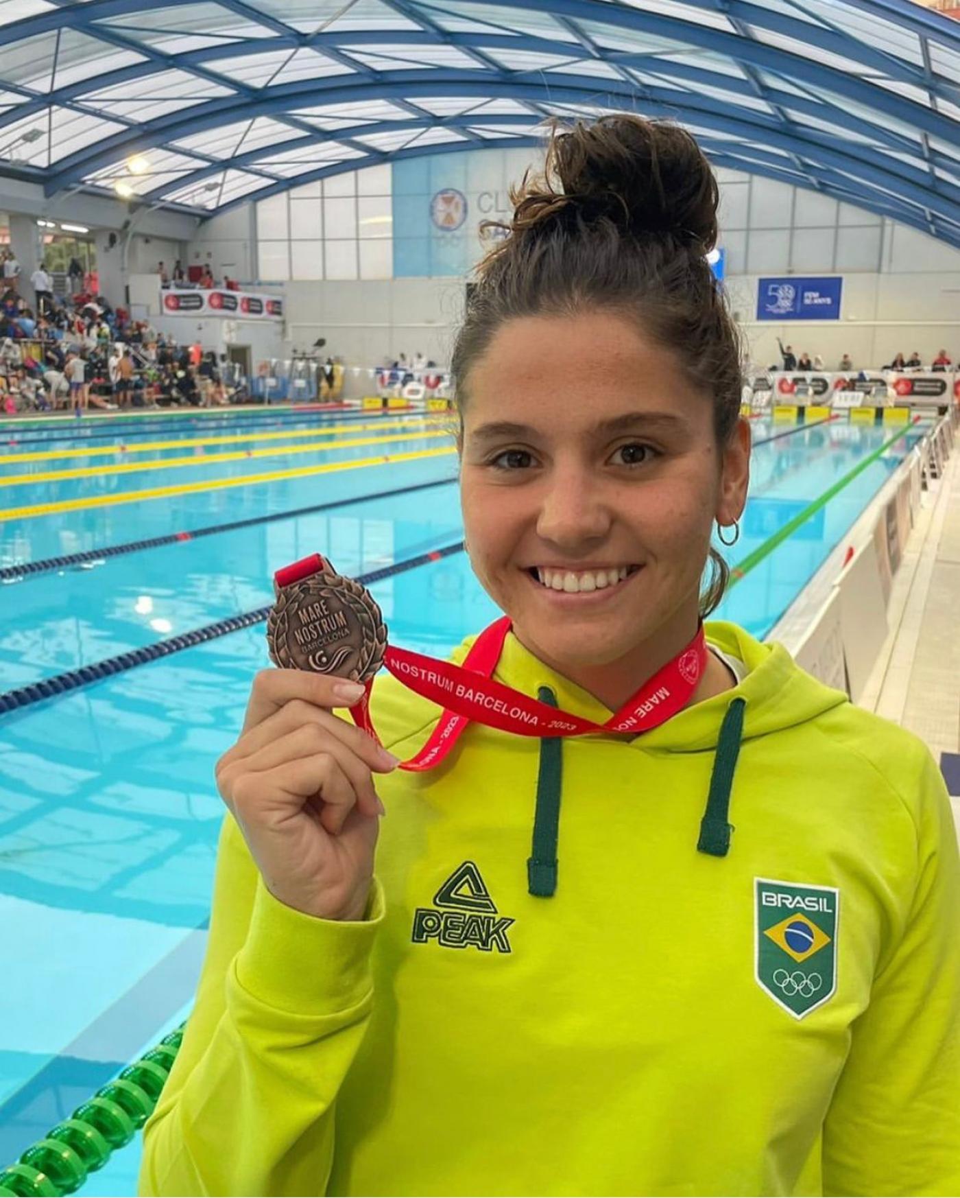 Beatriz Dizotti posa para foto segurando medalha e de costas para uma piscina. #paratodosverem