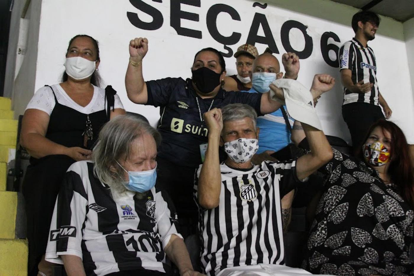 Rubens e José Roberto estão sentados lado a lado em cadeiras para assistir ao jogo. eles comemoram e sorriem com o gol marcado. Outras pessoas sentadas também comemoram. #paratodosverem