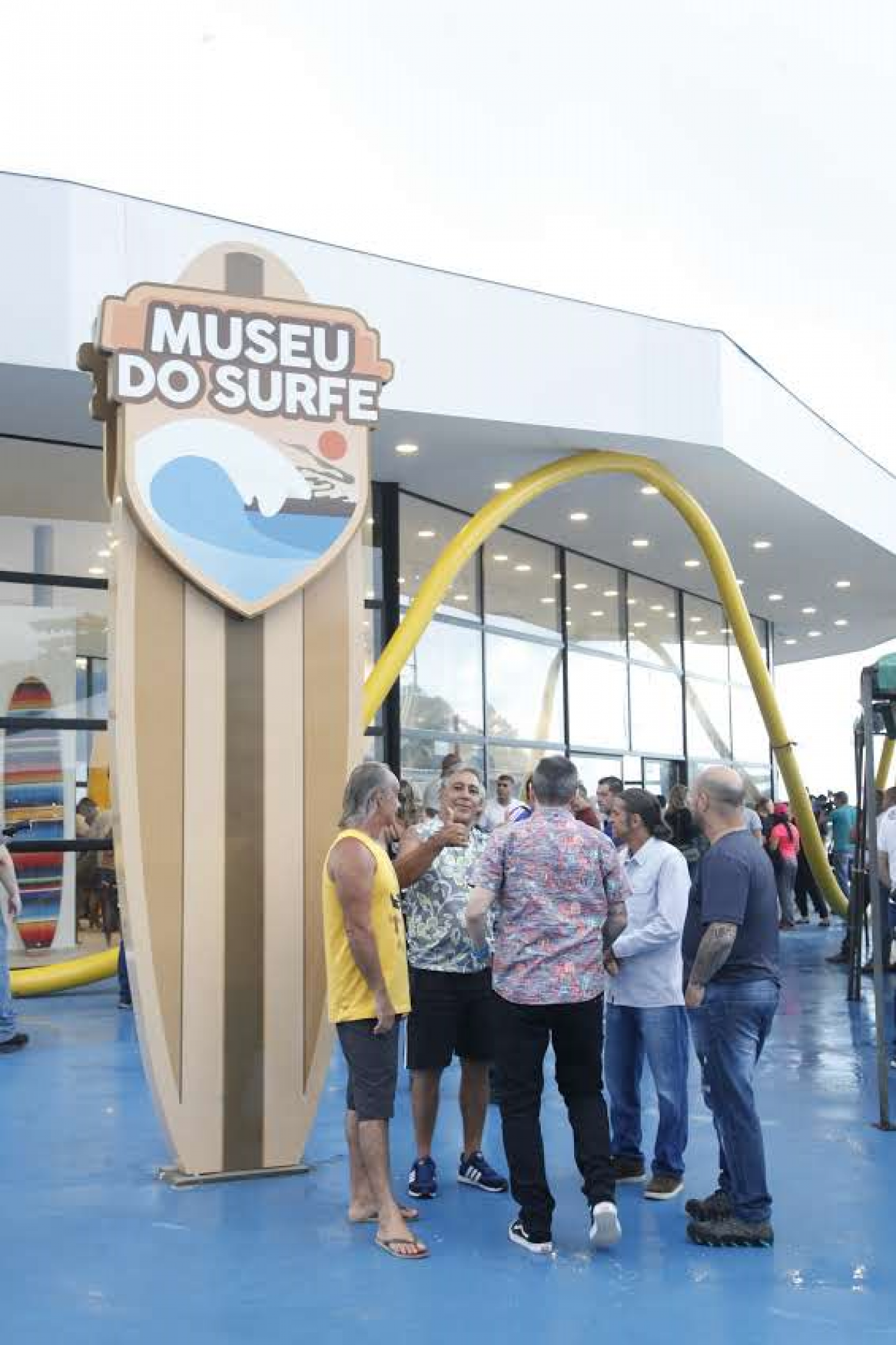 Totem em formato de prancha na entrada de museu onde se lê Museu do Surfe. Pessoas estão na frente conversando. #Pracegover