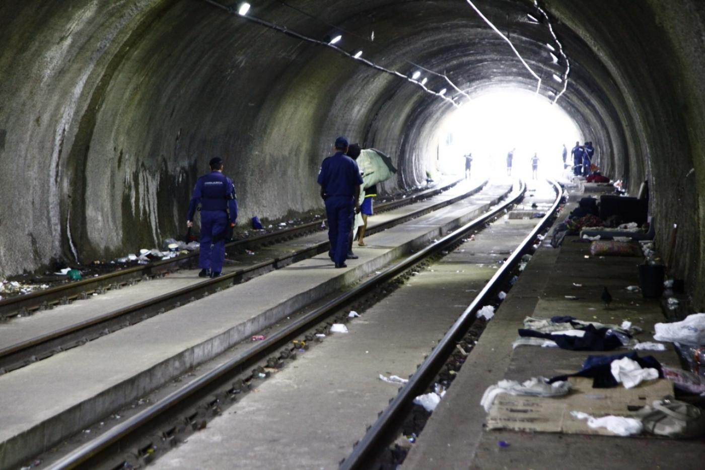 Dois guardas municipais estão dentro de túnel. Há uma pessoa à frente deles carregando um colchão. Nas laterais do túnel há peças de roupas no chão. #Pracegover 