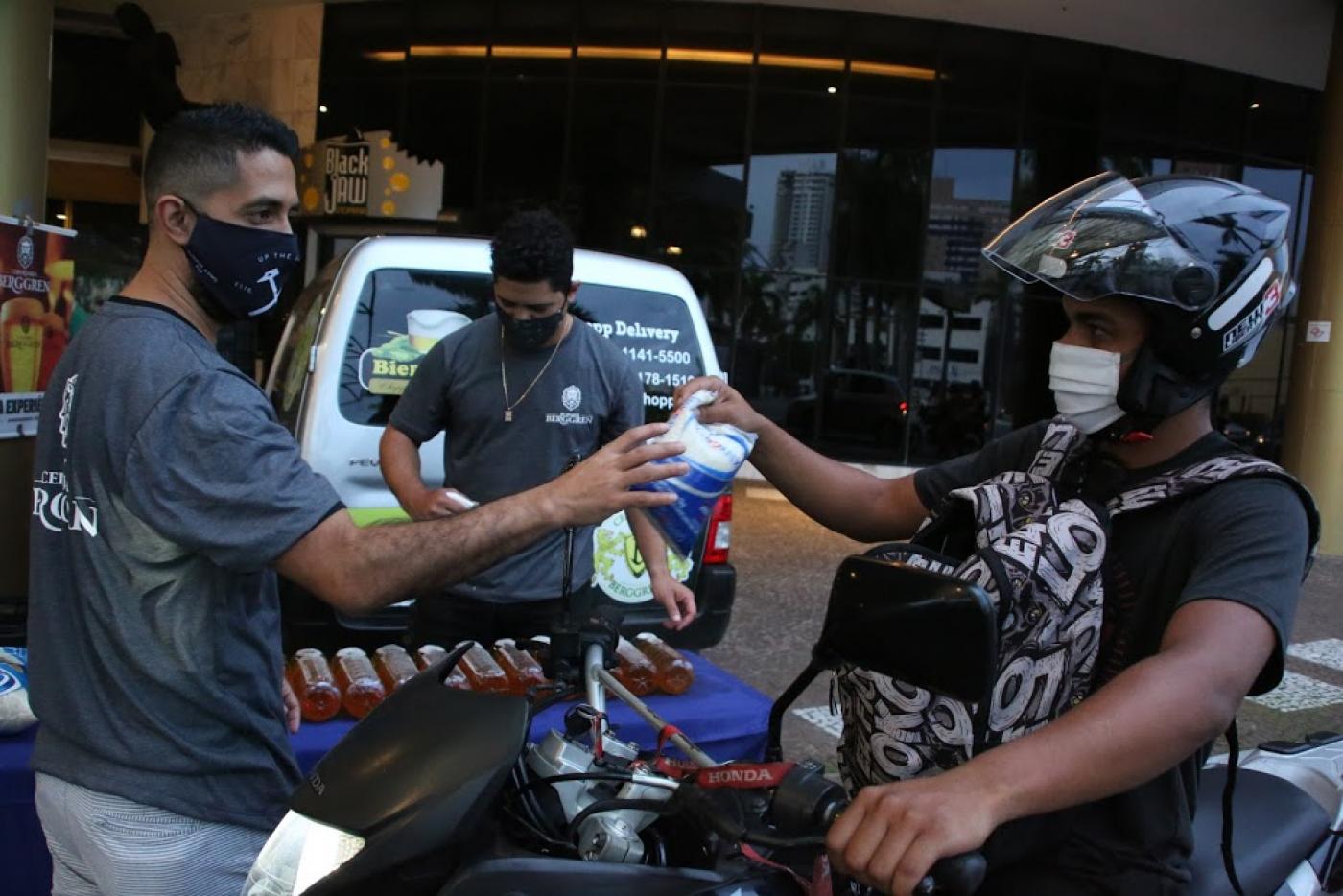 Homem sentado em moto, com capacete e máscara, entrega alimento para homem em pé, também de máscara. #pracegover