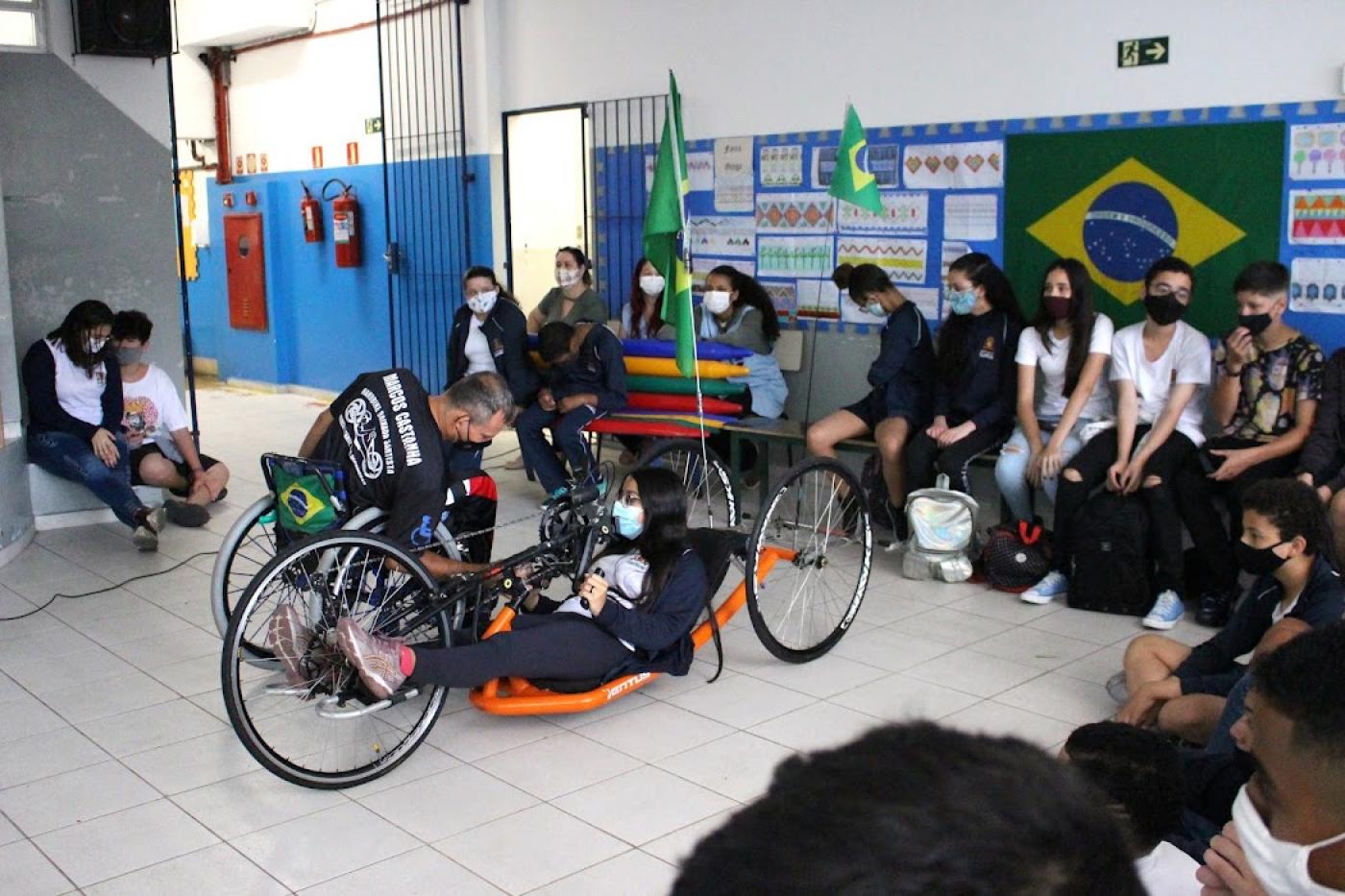 Paratleta mostra cadeira de roda para estudantes e outros observam #paratodosverem
