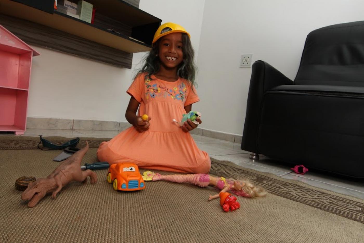 Luiza se diverte com brinquedos sobre tapete em uma sala. Há um sofá ao lado. #paratodosverem
