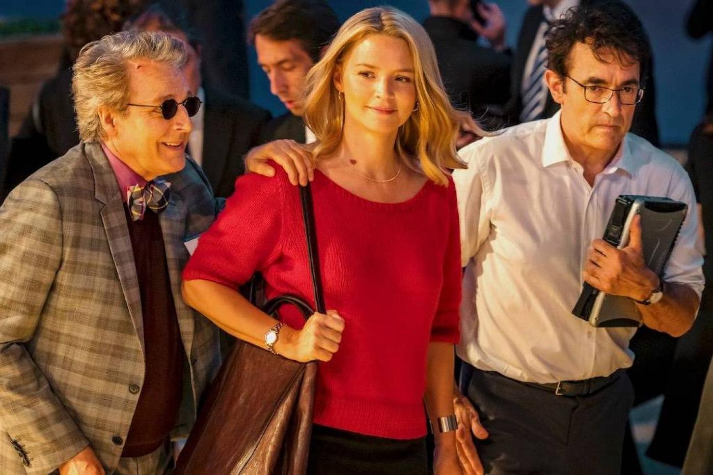 Cena de Adeus, Idiotas, com mulher no meio de dois homens. Ele estão caminhando. O homem da direita apoia a mão no ombro da mulher. #paratodosverem 
