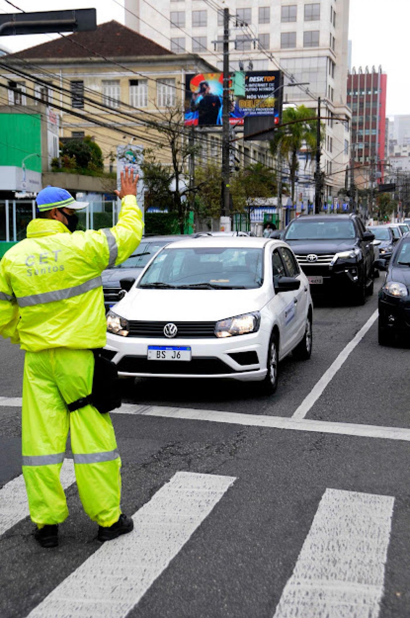 Agente uniformizado com a mão levantada orientando o trânsito. #pracegover