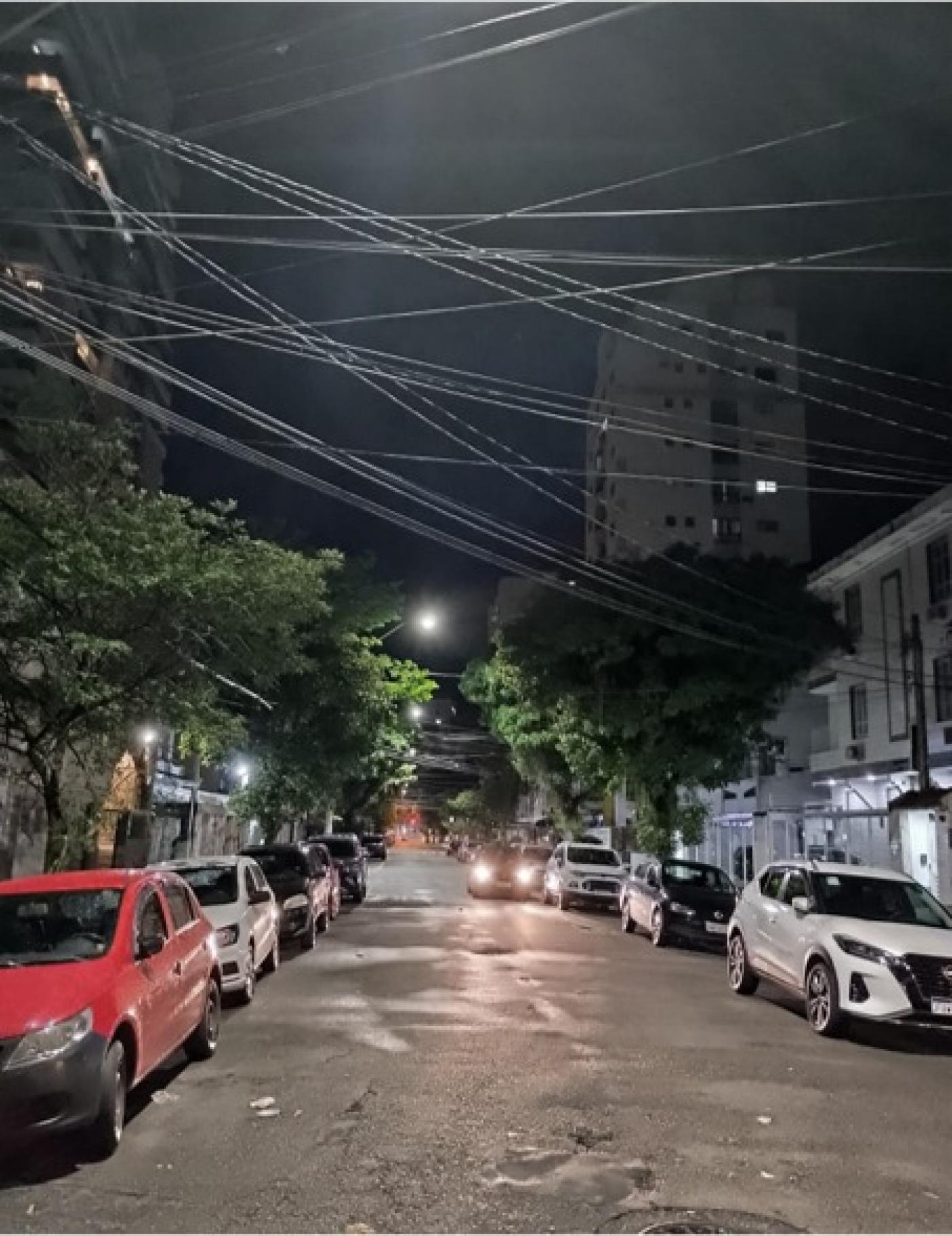 rua iluminada à noite. há carros estacionados. #paratodosverem 