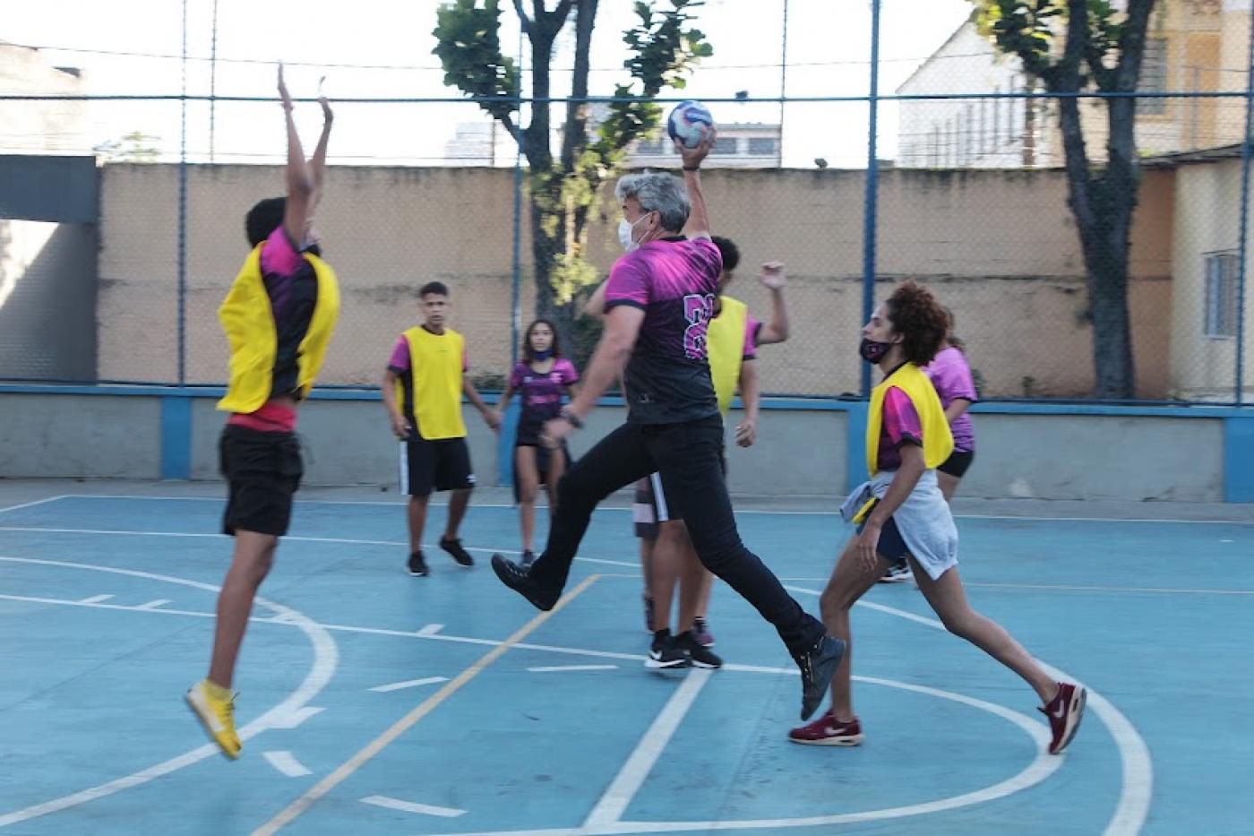 alexandre borgues está na quadra jogando basquete com adolescentes ele está segurando a bola e faz movimento de lance. Ele está de lado para a foto e seus dois pés estão fora do chão. #paratodosverem