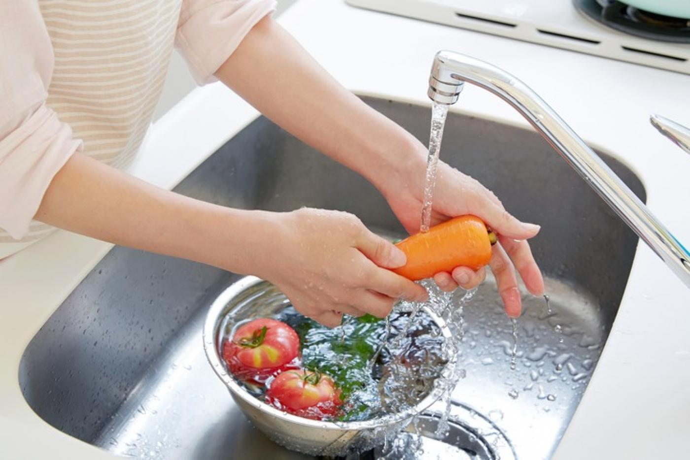 Mãos estão lavando cenoura sob jato de torneira. Abaixo há uma vasilha cheia d'água com tomates. #Paratodosverem