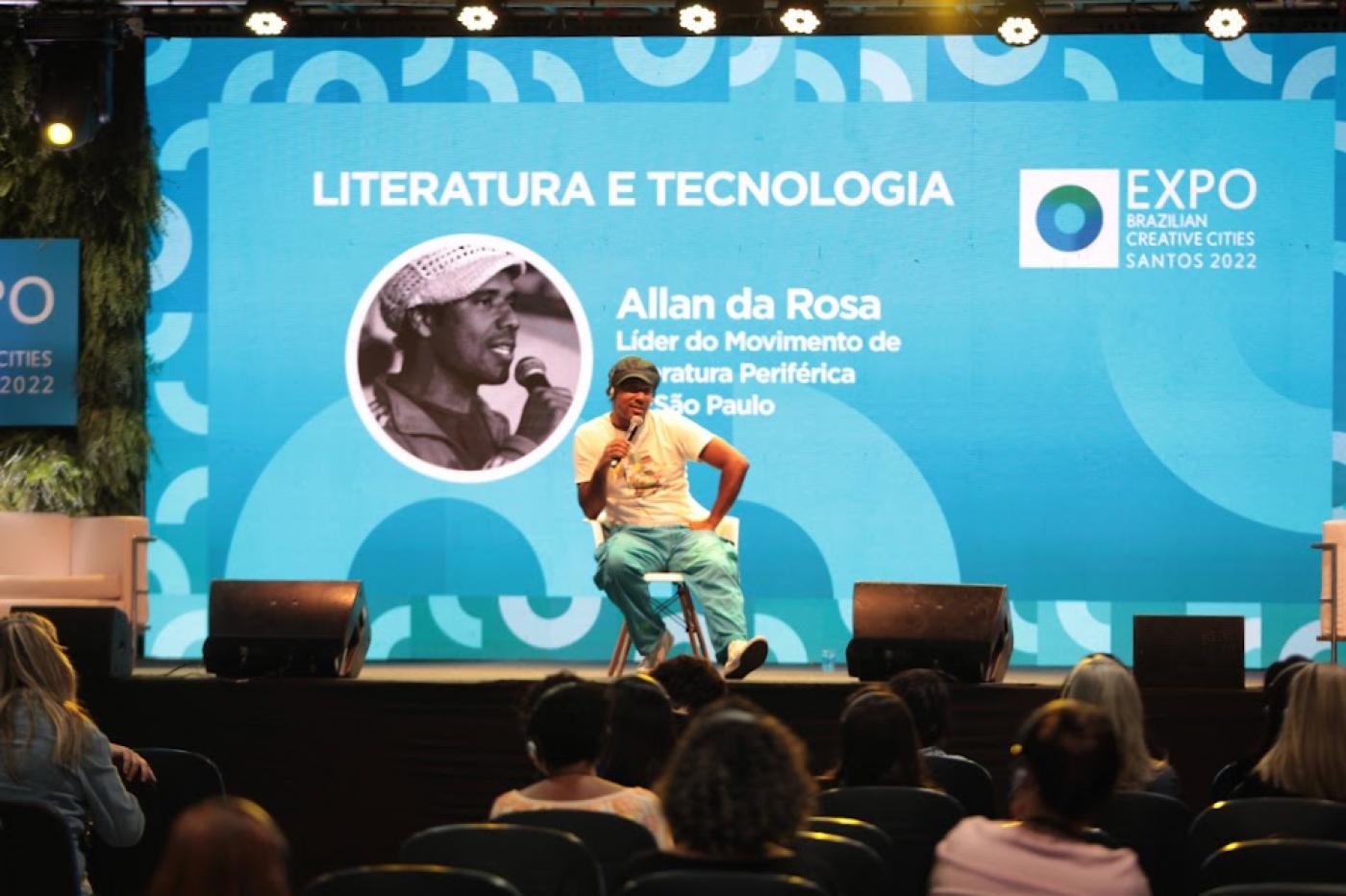Allan da Rosa fala do microfone sentado. #paratodosverem