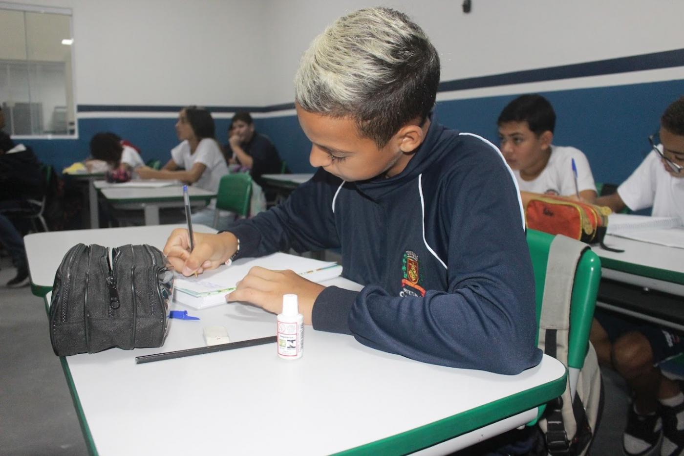 aluno está sentado em carteira escolar escrevendo em caderno. Há outros estudantes ao fundo. #paratodosverem 