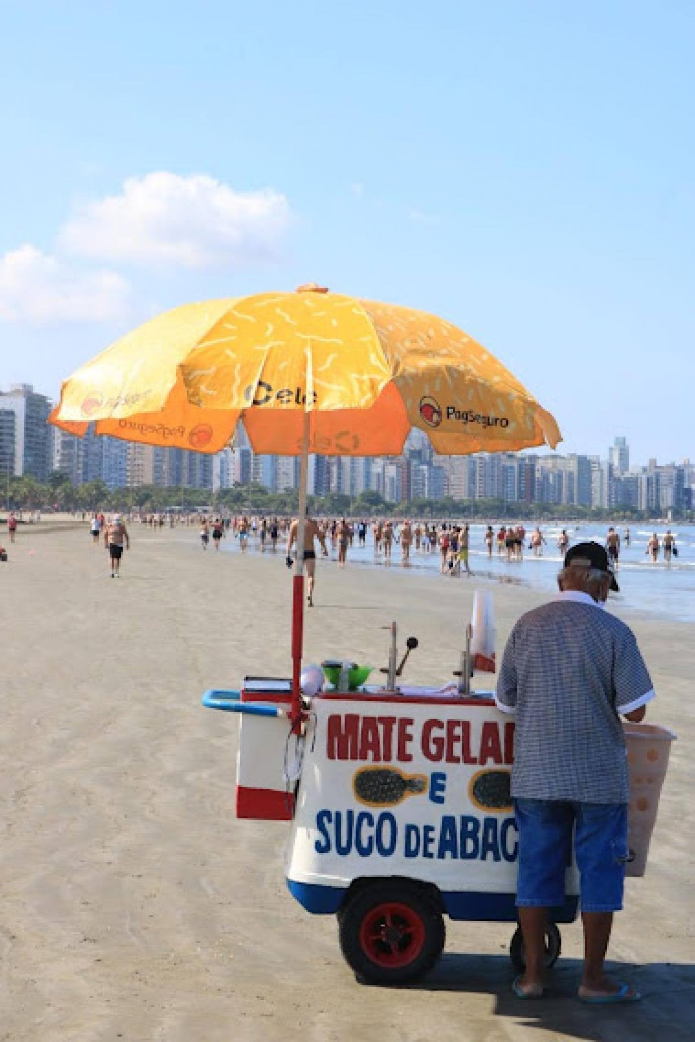 carrinho de mate na faixa de areia com guarda sol aberto. De costas para a foto está o ambulante. #paratodosverem