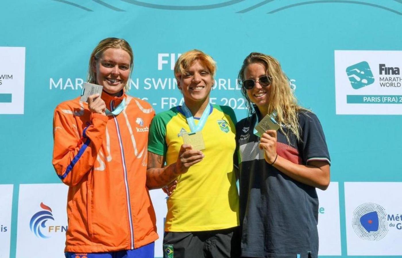 Ana Marcela e outras duas atletas exibem medalhas no pódio da prova. #pratodosverem