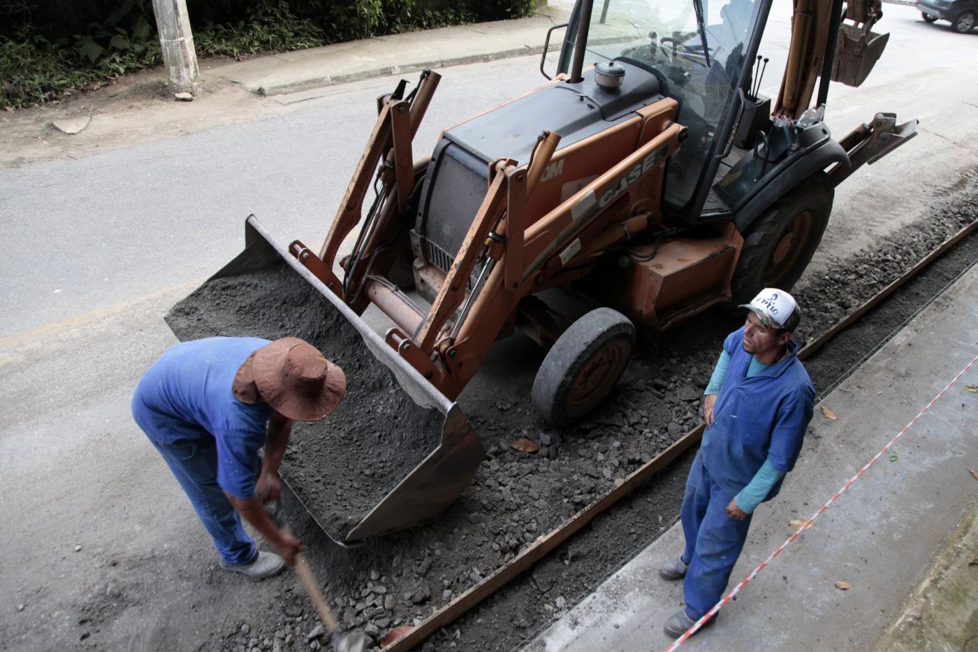 Dois operários trabalham ao lado de escavadeira #pracegover