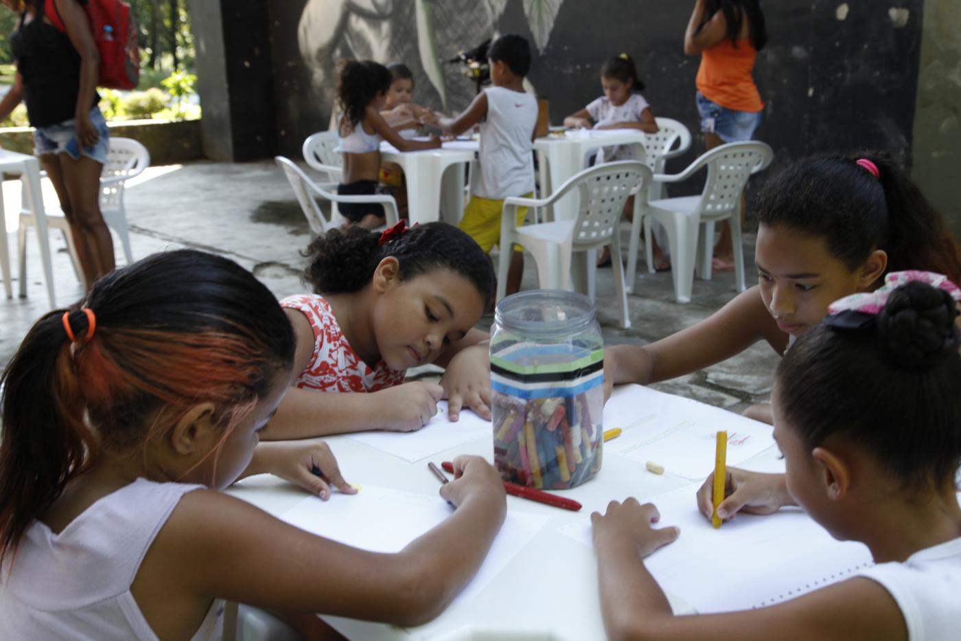 Crianças estão sentadas em torno de mesa. Elas desenham e pintam em folhas de papel. #Pracegover