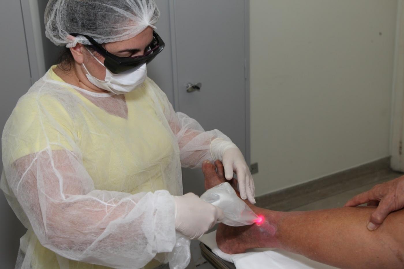 Mulher paramentada com avental, touca, luvas e óculos de proteção aplica laser na perna de um indivíduo. #Paratodosverem