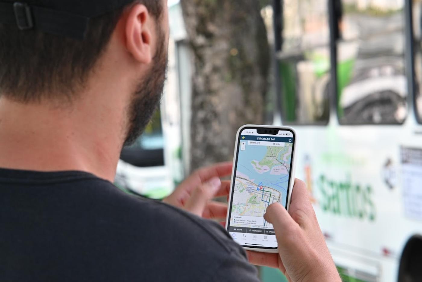 rapaz olha aplicativo com ônibus ao fundo #paratodosverem 