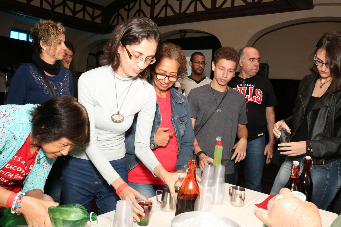 Oficina de drink à base de café. Participantes estão em pé em torno de uma mesa. Eles mexem em copos e garrafas. #Pracegover