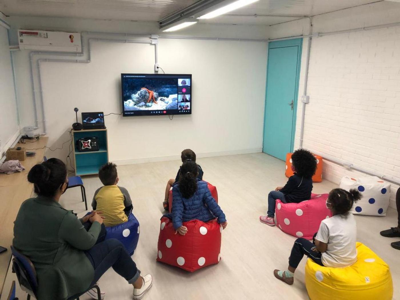 Crianças estão sentadas em pufs assistindo a v[ideo em tela ao fundo de sala. #paratodosverem