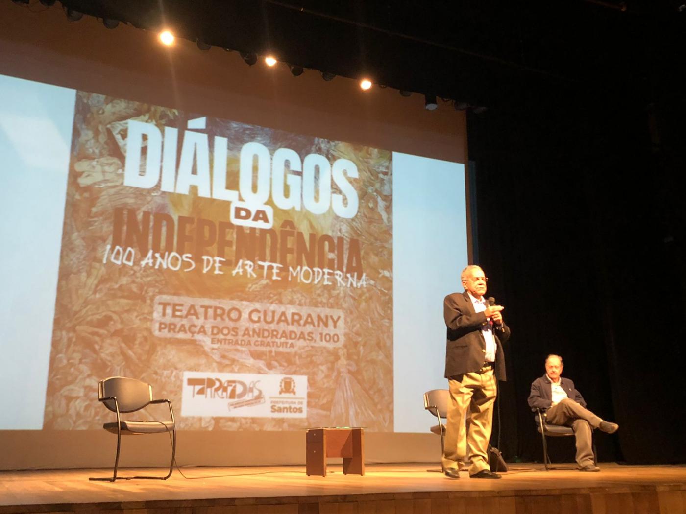 Dois homens no palco estão falando. Ao fundo há um painel no telão onde se lê Diálogos da Independência